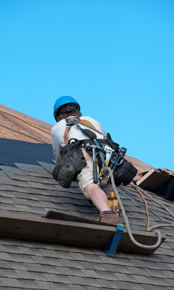 A worker in a blue helmet is climbing a sloped roof, secured with safety harnesses and tools.