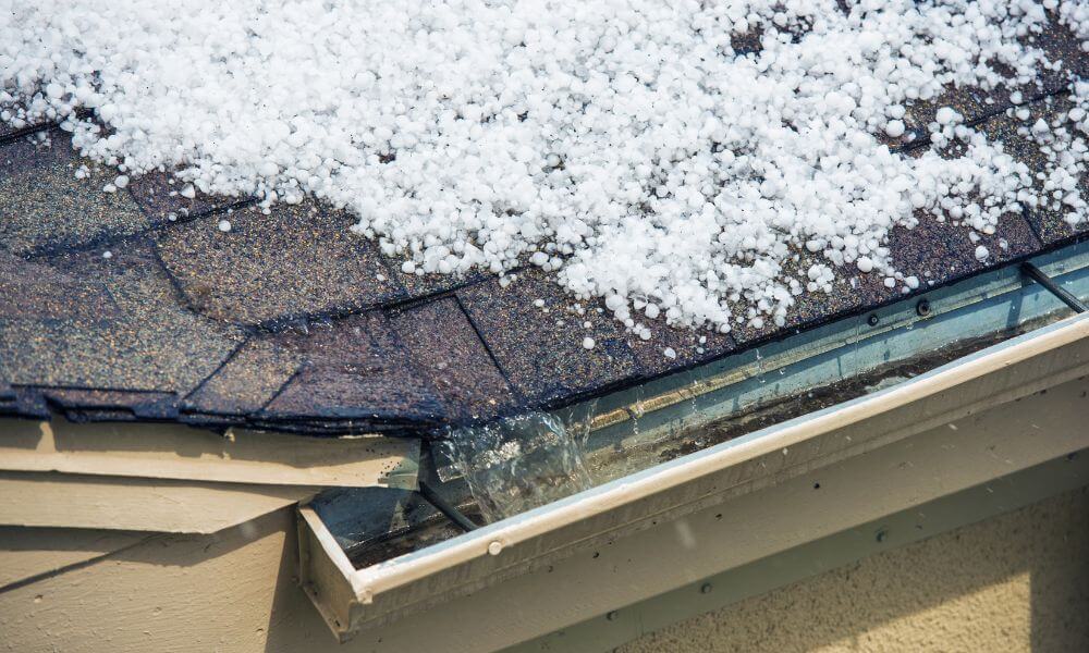 Hailstones covering a dark shingled roof, with water runoff in the gutter.
