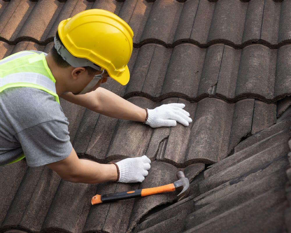 A worker in a yellow hard hat and safety vest is repairing a tiled roof using gloves and a hammer.