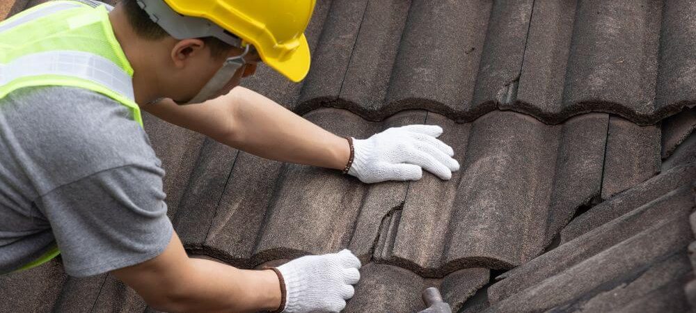 A worker in a yellow hard hat and safety vest is repairing a tiled roof using gloves and a hammer.